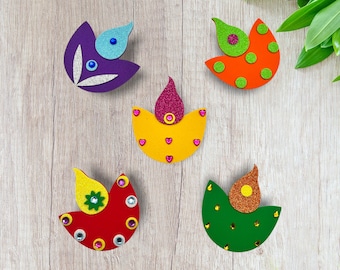 May include: Five colorful, decorative diya lamps. Each lamp features a unique design with glitter accents and jewel embellishments. The lamps are in shades of purple, orange, yellow, red, and green, with varying shapes and patterns.