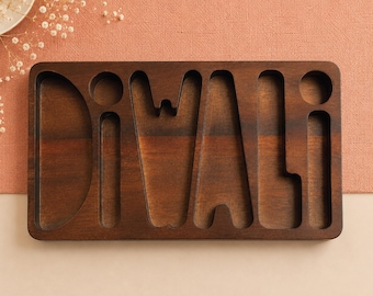 May include: A rectangular dark brown wooden tray featuring carved cutouts that spell out the word DIVALI. The tray has a smooth, polished finish with visible wood grain patterns throughout the surface, designed with distinct letter-shaped compartments.
