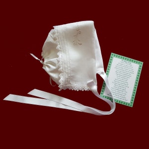 May include: White linen baby bonnet with lace trim and a white satin ribbon. The bonnet has an embroidered shamrock design and the text "The Magic Words" is printed on a green card with shamrock accents.