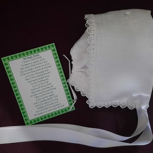 May include: A white baby bonnet with lace trim and a white satin ribbon. The bonnet is embroidered with a floral design. A card with a poem about the bonnet is included. The poem is titled "The Magic Hanky".