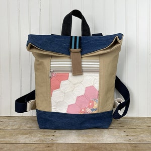 May include: A beige and blue canvas backpack with a patchwork quilt design on the front pocket. The backpack has a roll-top closure and a black strap.