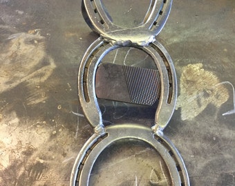 Può includere: Un supporto in metallo realizzato con tre ferri di cavallo saldati insieme. Il supporto è di colore argento e ha un aspetto rustico.