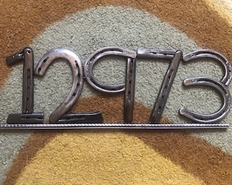 May include: A metal house number made from horseshoes, the number is 1273. The horseshoes are arranged in a row on a metal bar.