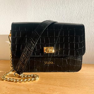 May include: Black crossbody bag with a crocodile embossed leather texture and a gold chain strap. The bag has a gold clasp and the brand name "VILNA" is embossed on the front.