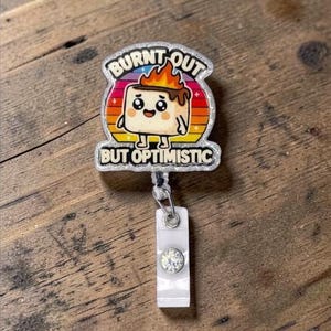 May include: A badge reel with a cartoon marshmallow character with flames on top. The text on the badge reel says "BURNT OUT" above the character and "BUT OPTIMISTIC" below. The reel has a clear plastic holder with a rhinestone.