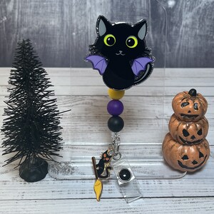 May include: A black cat with purple wings and yellow eyes is on a black and purple beaded lanyard with a black and gold witch's broom charm. The lanyard is attached to a clear plastic badge holder. A stack of three orange pumpkins with jack-o-lantern faces is in the background.