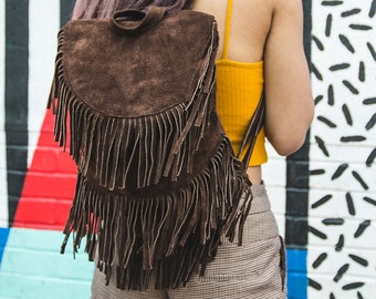May include: A brown suede backpack with fringe detailing. The backpack is worn by a person with purple hair and a brown hat. The person is standing in front of a colorful wall with geometric patterns.