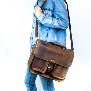 May include: A woman wearing a blue denim jacket carries a brown leather satchel bag with a long strap over her shoulder.
