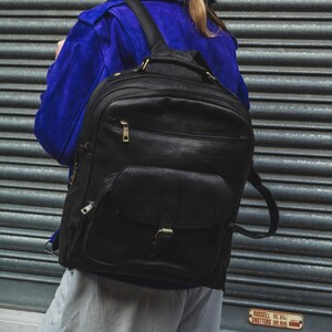 May include: A person wearing a blue jacket and a black leather backpack. The backpack has a large front pocket with a buckle closure.