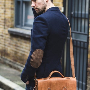 Op de afbeelding: Een man draagt een donkerblauw blazer met een bruine leren patch op de elleboog en draagt een bruine leren schoudertas met een klep en een lange band.