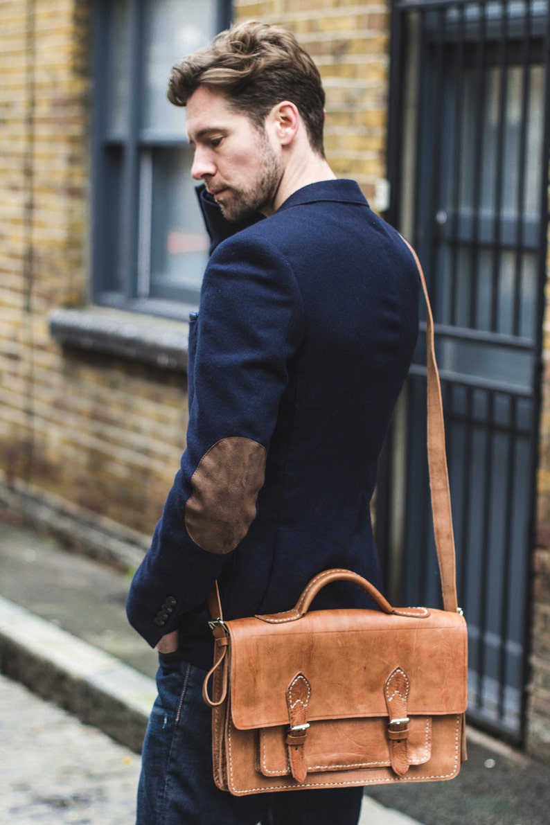 Op de afbeelding: Een man draagt een donkerblauw blazer met een bruine leren patch op de elleboog en draagt een bruine leren schoudertas met een klep en een lange band.