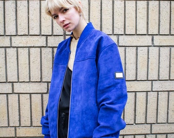 May include: A woman wearing a blue suede bomber jacket with a zip fastening and a small white label on the sleeve. She is standing in front of a brick wall.