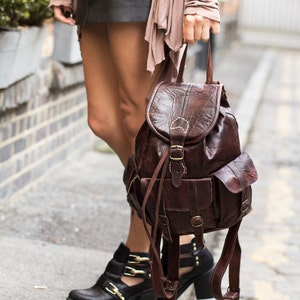 May include: A brown leather backpack with multiple pockets and adjustable straps. The backpack is worn by a person wearing a black leather skirt and a light brown cardigan.