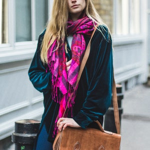 Op de afbeelding: Een vrouw draagt een teal fluwelen jasje met een roze en paarse sjaal en een bruine leren schoudertas.