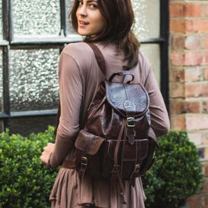 May include: A woman wearing a brown shirt and a brown leather backpack with a flap and buckle closure. She is standing in front of a window and a brick wall.