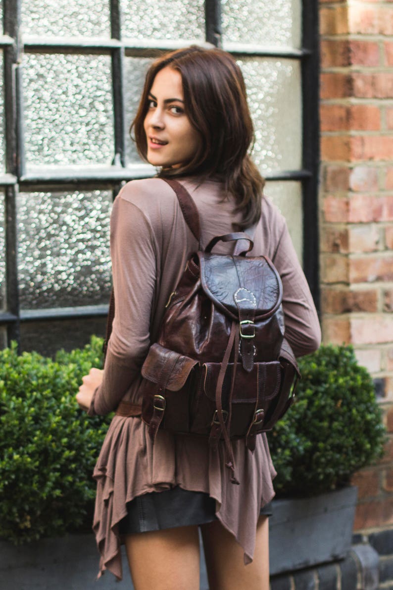 May include: A woman wearing a brown shirt and a brown leather backpack with a flap and buckle closure. She is standing in front of a window and a brick wall.