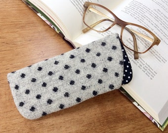 May include: A white and black polka dot glasses case made of burlap fabric. The case is lying on top of an open book with a pair of brown glasses on top of the book.