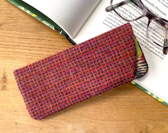 May include: A pink, green and black tweed glasses case with a floral pattern on the back. The case is lying on a wooden surface next to a book and glasses.