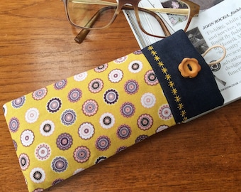 May include: A yellow and purple patterned glasses case with a navy blue flap and a brown button. The case is lying on a wooden surface.
