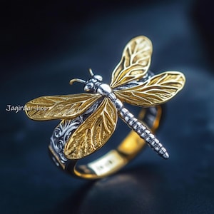 May include: A silver ring featuring a gold dragonfly design. The dragonfly's wings are detailed and intricate, making this ring a statement piece.