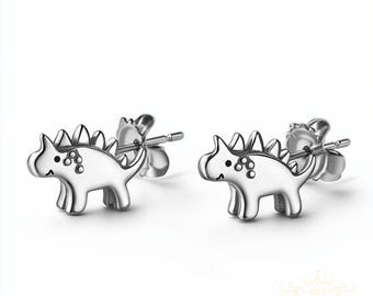 May include: A pair of silver stud earrings featuring a cartoon dinosaur design. The dinosaurs are facing forward with their mouths open in a smile.