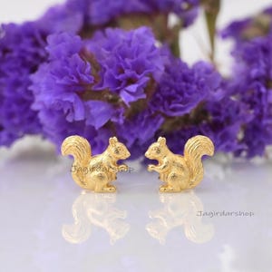 May include: A pair of gold-toned squirrel-shaped stud earrings with textured fur detailing. The earrings are positioned side-by-side, each featuring a bushy tail and a small ear. The text Jagirdarshop is visible near the squirrel figures.