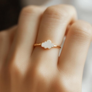 Puede incluir: Un anillo dorado con un diseño en forma de nube blanca. El diseño del anillo es simple y elegante, con la nube como foco central. Una joya delicada, perfecta para el uso diario. La nube es blanca.