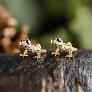 Puede incluir: Un par de pendientes de lagarto de color dorado con ojos negros. Los lagartos están mirando hacia adelante con la boca cerrada.