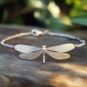 Könnte beinhalten: Ein silbernes Armband mit einem Libellen-Anhänger. Die Libelle ist mit filigranen Flügelmustern verziert.