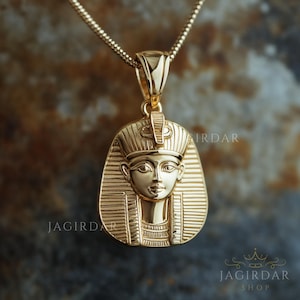 Könnte beinhalten: Eine goldene Halskette mit einem Anhänger, der eine detaillierte Darstellung eines ägyptischen Pharaos zeigt. Der Anhänger ist aufwendig gestaltet mit einer Krone und einem Gesicht mit einem ruhigen Ausdruck.
