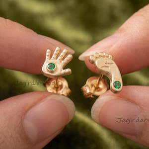 Puede incluir: Un par de pendientes de oro tipo botón con un diseño en forma de mano y otro en forma de pie. Cada pendiente está decorado con una pequeña piedra preciosa redonda de color verde engastada en el centro de la palma y el talón respectivamente.