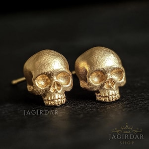 May include: Two gold-coloured skull stud earrings. The earrings feature eye sockets, a nose cavity, and teeth. They are set against a dark background. The brand name "JAGIRDAR SHOP" is visible in the lower right corner.