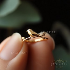 Könnte beinhalten: Ein goldfarbener Ring mit einem kleinen Vogel, der auf einem Zweig sitzt. Der Ring ist zart und filigran, perfekt für den täglichen Gebrauch.