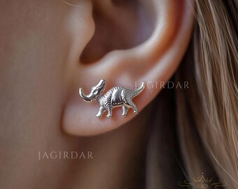 May include: A silver-colored dinosaur stud earring is shown on an ear. The earring is a detailed depiction of a dinosaur, with textured skin and a curved horn. The earring is small and delicate, suitable for everyday wear.