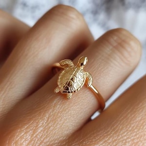 Op de afbeelding: Een goudkleurige ring met een gedetailleerd schildpadontwerp. De schildpad heeft een getextureerd schild en is geplaatst op een dunne, gouden band. De ring wordt op een vinger getoond, wat het ontwerp en de grootte van de sieraden benadrukt.