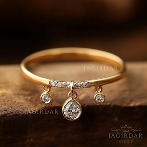 May include: A gold ring with three dangling diamond pendants. The ring band is thin and polished, with a row of small diamonds set into the top. The pendants are pear and round cut diamonds. The ring is on a wooden surface.
