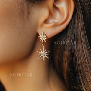 May include: Gold star-shaped earrings with clear crystals. The earrings are dangling and have two stars on each earring.