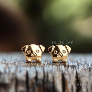May include: A pair of gold-toned stud earrings featuring a small, detailed design of a dog. The earrings are on a wooden surface.
