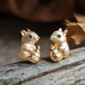 May include: A pair of gold-toned metal stud earrings shaped like small mice, each holding a textured acorn. The mice have detailed fur markings and small black eyes. The earrings are positioned side by side, showcasing the metallic finish.