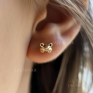 May include: Gold frog stud earrings. The earrings are small and delicate, and the frog is facing forward with its mouth open.