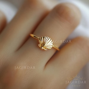 May include: A gold-toned ring featuring a small, detailed seashell charm centered on a thin, polished band. The seashell design includes vertical ridges and a scalloped edge. The text JAGIRDAR is visible on the left and right sides.