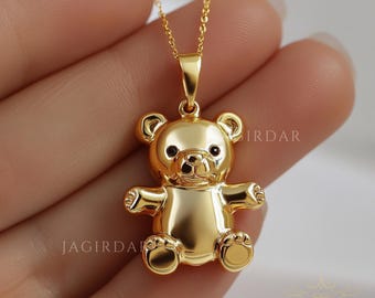 May include: A gold-tone necklace with a teddy bear pendant. The bear is sitting upright with its arms outstretched. The necklace is on a hand.