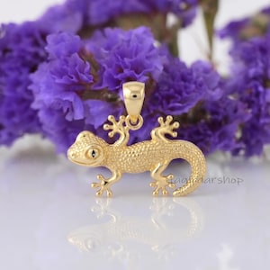 May include: A gold-toned lizard pendant featuring a textured, granular surface, a curved tail, and a small bail at the top for attachment. The lizard is depicted in a horizontal position with its four legs extended, showcasing a detailed head.