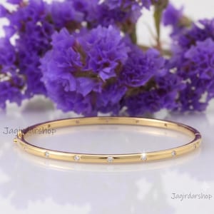 Op de afbeelding: Een dunne gouden armband met een reeks kleine, gelijkmatig verdeelde ronde heldere stenen langs de buitenkant. De armband heeft een scharniersluiting en is horizontaal geplaatst tegen een achtergrond van paarse bloemen.