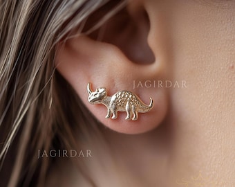 May include: Gold dinosaur earrings. The earrings are shaped like a small dinosaur with a textured surface. The earrings are shown on a person's ear.