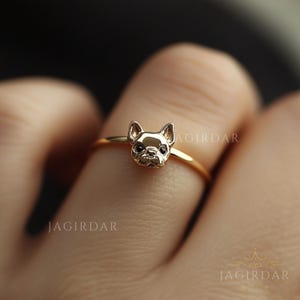 May include: A gold-colored ring featuring a detailed French Bulldog head. The dog's face has black accents for the eyes and nose. The ring is delicate and sits on a finger, showcasing the jewelry's design.