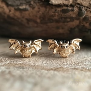 Può includere: Un paio di orecchini a forma di pipistrello dorati con occhi neri. Gli orecchini sono su una superficie di legno.