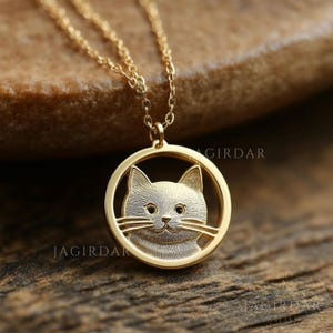 Puede incluir: Un collar dorado con un colgante que representa la cara de un gato dentro de un marco circular. El gato tiene detalles, incluyendo bigotes y una superficie texturizada. El collar se muestra sobre una superficie de madera.