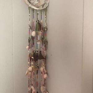 May include: A white seashell wind chime with multiple strands of colorful beads, shells, and bells. The chime is hanging from a white wall.