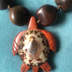 May include: A handcrafted necklace featuring a wooden turtle pendant with a shell inlay. The pendant is a reddish-brown color, and the necklace has large, dark brown beads. The necklace is strung on a brown cord.